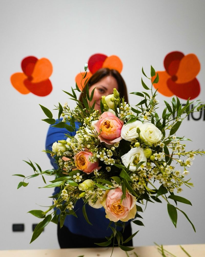 Margareta porge verso l'obiettivo un bouquet di rose pesca, lisianthus bianchi e fiori di cera, davanti a una parete con papaveri dipinti