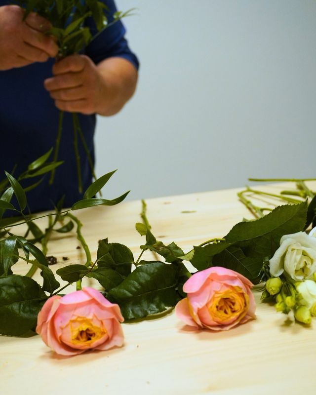 Mani che raccolgono rami verdi sopra un tavolo di legno con due rose rosa e boccioli bianchi